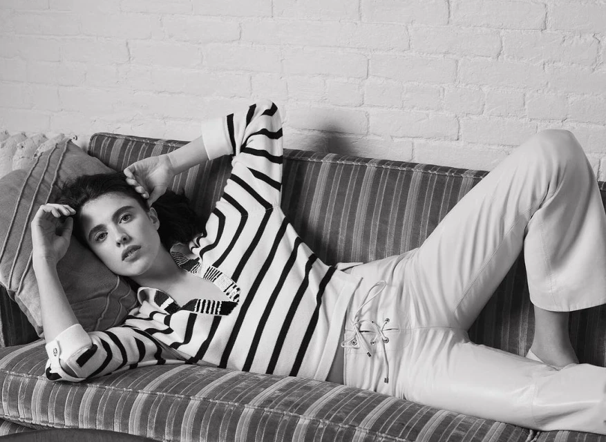 A young woman with dark hair lies on a striped couch wearing a striped shirt and white pants, looking at the camera against a brick wall.