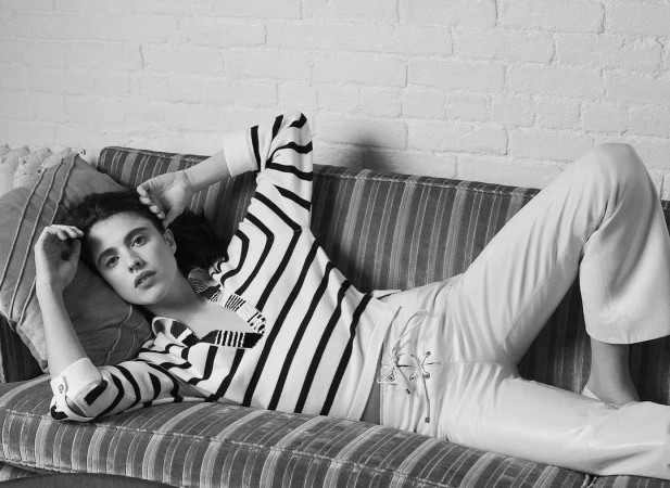 A young woman with dark hair lies on a striped couch wearing a striped shirt and white pants, looking at the camera against a brick wall.