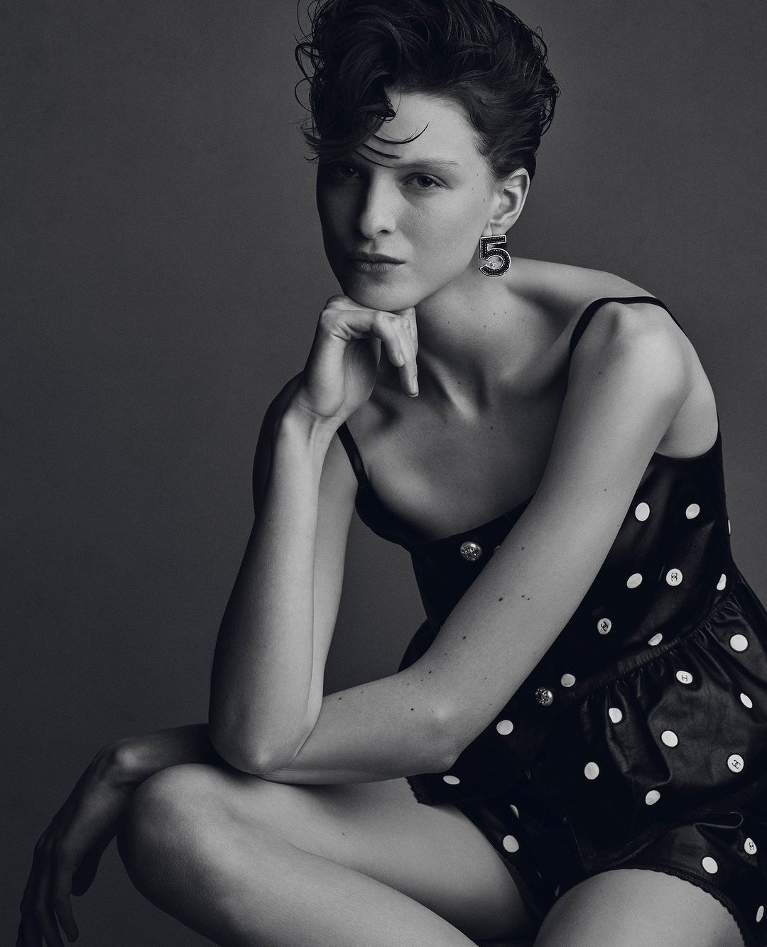 Studio portrait of a young adult woman with dark hair in a black polka-dot dress, gazing at the camera, against a grey background.