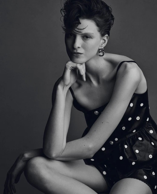 Studio portrait of a young adult woman with dark hair in a black polka-dot dress, gazing at the camera, against a grey background.