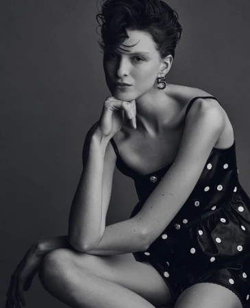 Studio portrait of a young adult woman with dark hair in a black polka-dot dress, gazing at the camera, against a grey background.