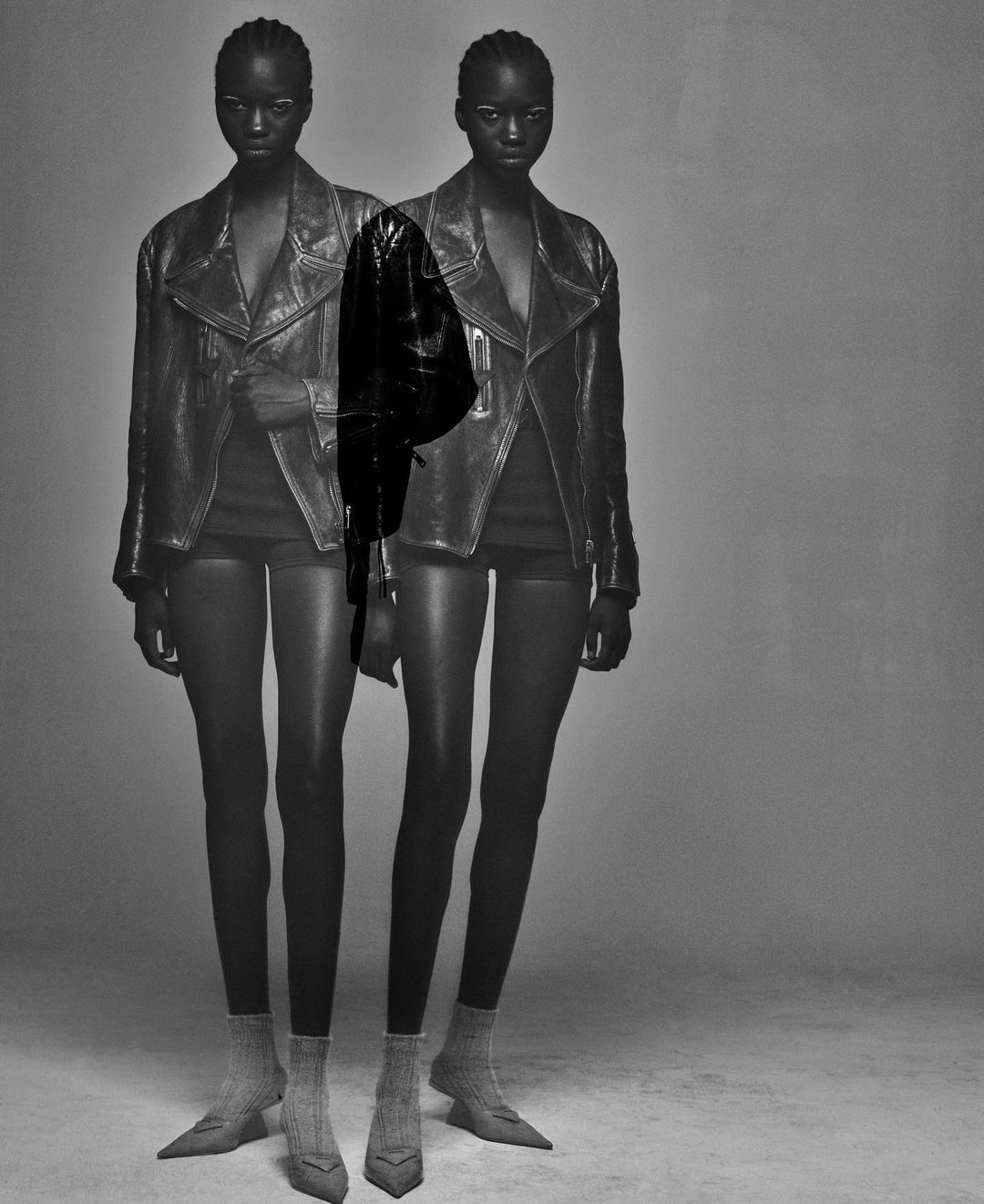 Two Black women stand in a studio wearing leather jackets, shorts, and pointy sock boots. A dark jacket is superimposed over the figures.