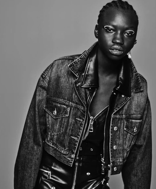 Black and white portrait of a woman with creative makeup, wearing a denim jacket and leather outfit, against a neutral gray backdrop.