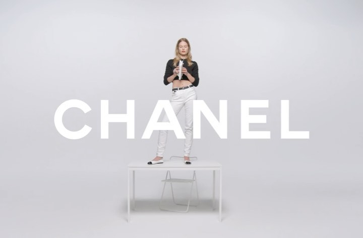 A woman in a black crop top and white pants stands on a white table against a white background, with the word CHANEL in large white letters.