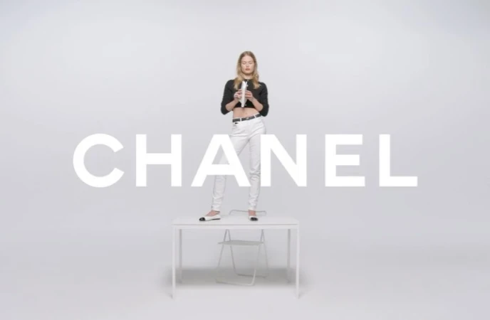 A woman in a black crop top and white pants stands on a white table against a white background, with the word CHANEL in large white letters.