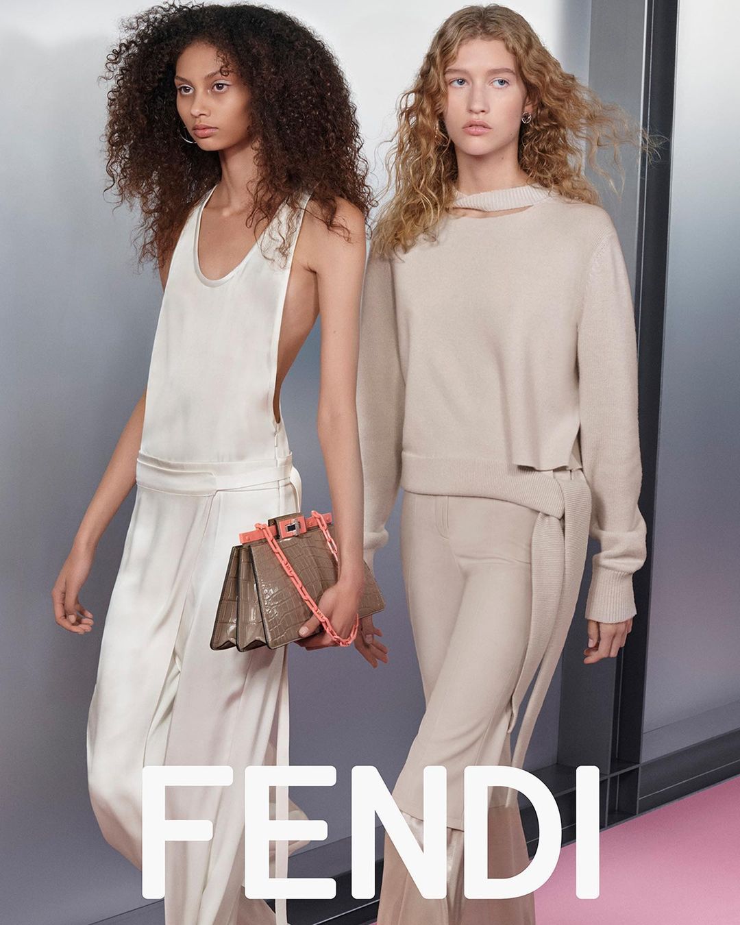 Two fashion models showcase beige outfits and an elegant Fendi handbag on a soft pink runway with modern metallic wall decor.