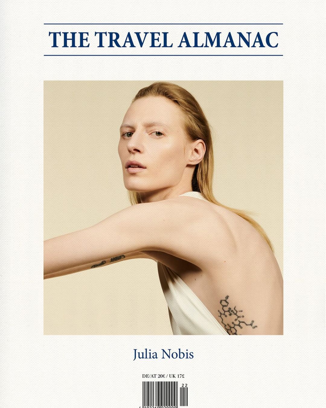 Blonde woman in white dress posing in front of a beige background, showcasing a geometric tattoo and the cover of a travel almanac.