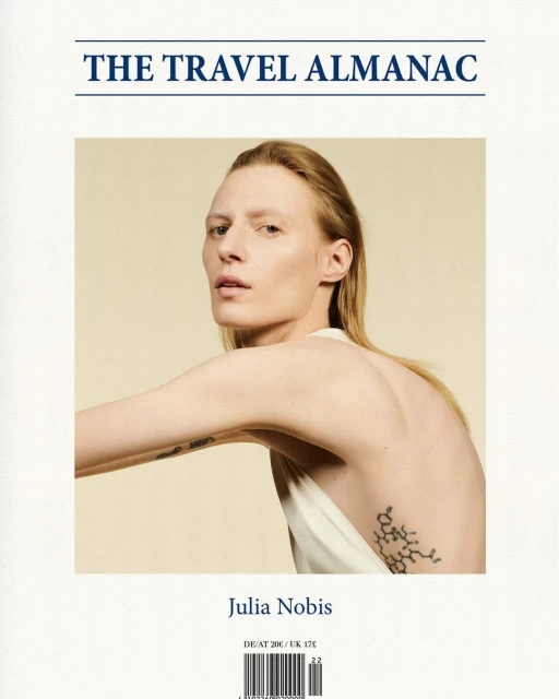 Blonde woman in white dress posing in front of a beige background, showcasing a geometric tattoo and the cover of a travel almanac.