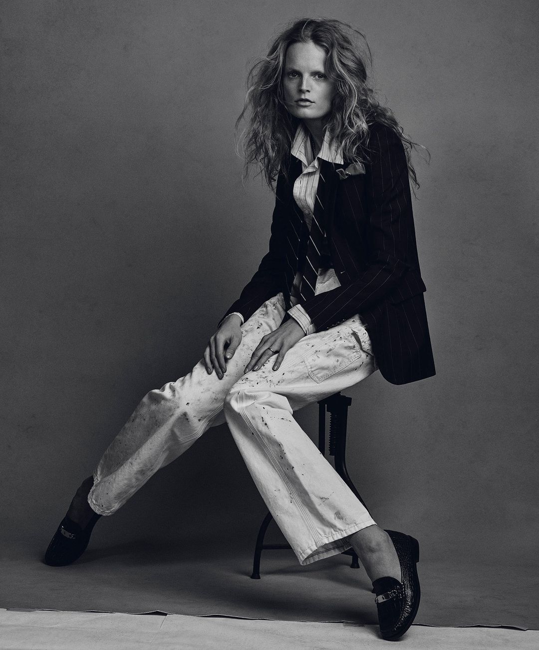 Model seated on a stool wearing a dark pinstripe blazer, white collared shirt, and paint-splattered white trousers against a grey backdrop.