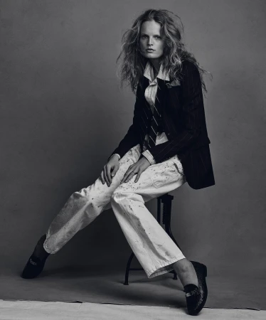 Model seated on a stool wearing a dark pinstripe blazer, white collared shirt, and paint-splattered white trousers against a grey backdrop.
