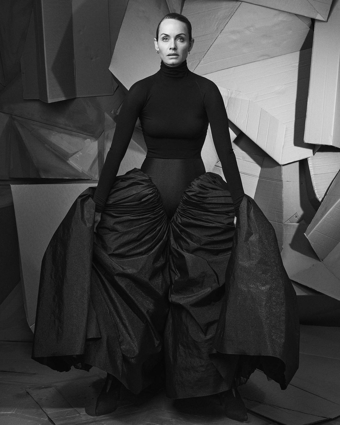Portrait of a woman in black clothing with an expressive skirt, posing in front of geometric panels in a monochromatic photo studio setting.