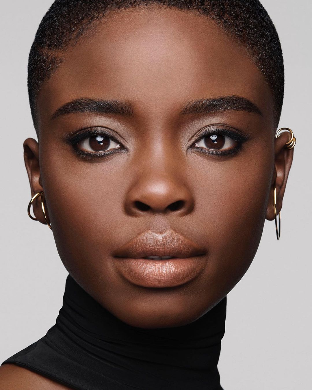 Close-up portrait of a young African woman with makeup wearing a black turtleneck, set against a neutral gray backdrop in a studio setting.