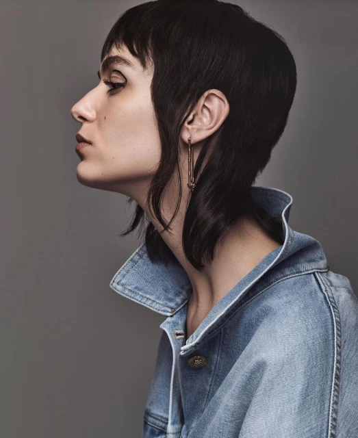 Profile shot of a stylish adult female with short dark hair and winged eyeliner wearing a light-wash denim jacket against a gray background.