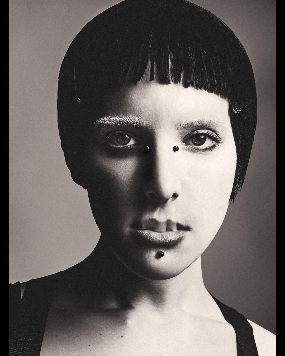 Striking monochrome portrait of a woman with a blunt black bob haircut and unique facial piercings in a bright studio setting.