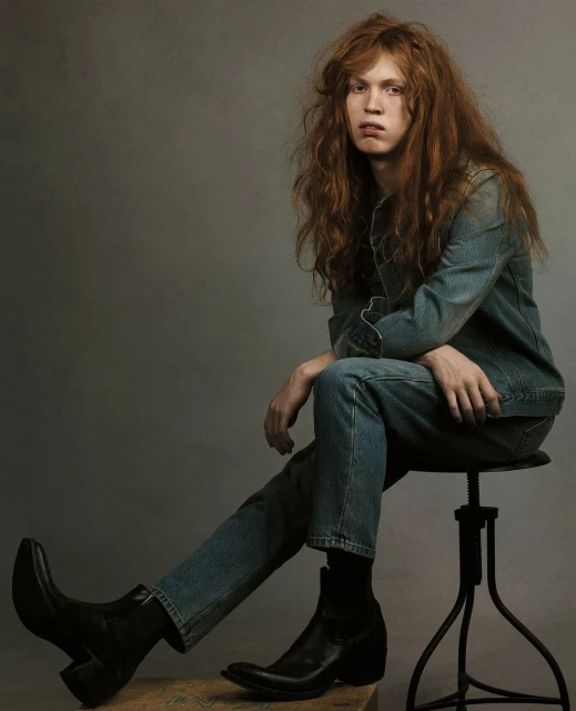 Portrait of a young adult with long red hair, sitting on a stool. The model is wearing denim clothing and black leather boots in a studio.