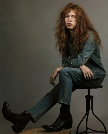 Portrait of a young adult with long red hair, sitting on a stool. The model is wearing denim clothing and black leather boots in a studio.
