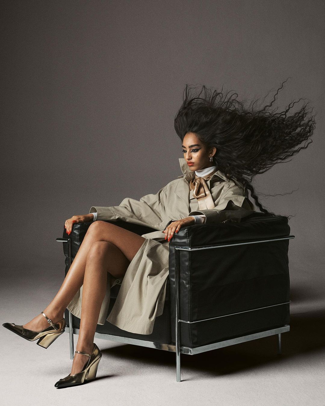 Elegant woman with windblown hair poses in a chair wearing a trench coat, top, metallic heels, and jewelry in a studio setting.