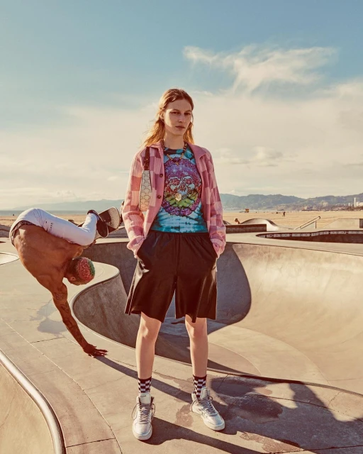 Woman in shorts and tie-dye shirt standing in a skatepark with a man doing a handstand on a skateboard in the background.