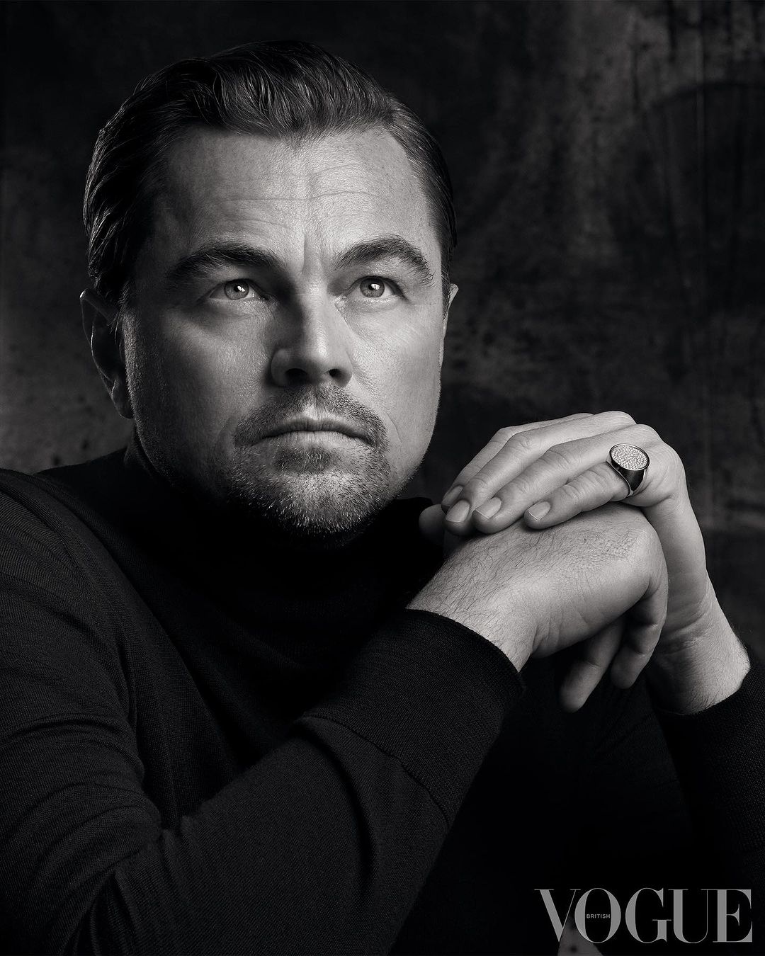 Monochrome portrait of Leonardo DiCaprio in a black turtleneck, hands folded, wearing a signet ring, against an abstract background.