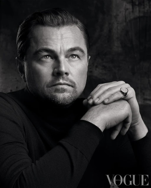 Monochrome portrait of Leonardo DiCaprio in a black turtleneck, hands folded, wearing a signet ring, against an abstract background.