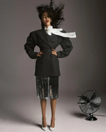 Stylish woman in a coat and skirt posing in a photo studio with wind blowing through her hair, and a vintage electric fan.