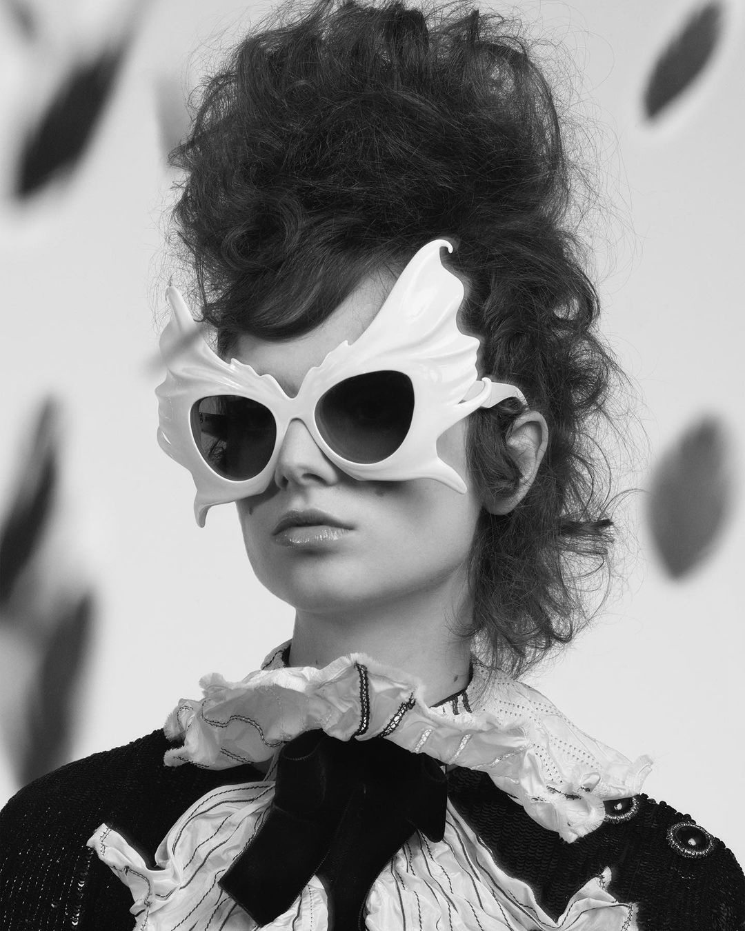 Model with voluminous updo hairstyle wearing sculptural white winged sunglasses and a ruffled collared blouse by Craig McDean.