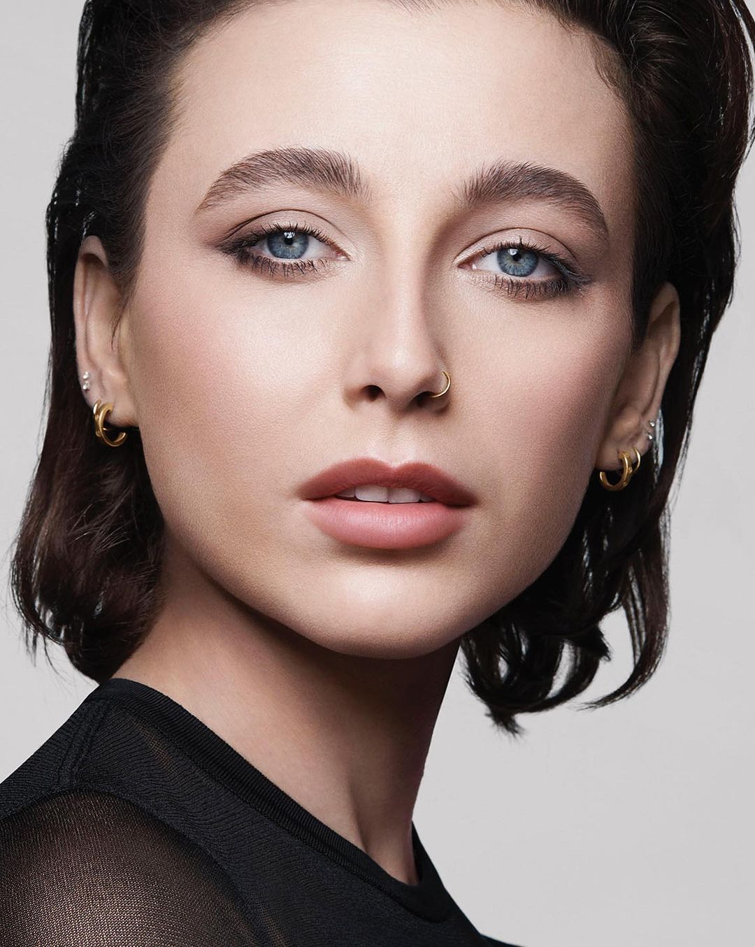 Close-up portrait of a woman with wet-look dark hair, blue eyes, and subtle makeup against a neutral grey background