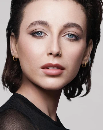 Close-up portrait of a woman with wet-look dark hair, blue eyes, and subtle makeup against a neutral grey background