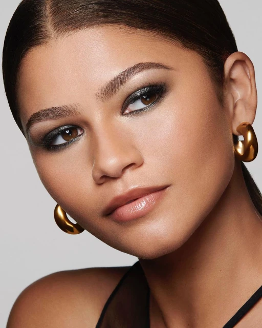 Close-up portrait of a woman with golden hoop earrings, smokey eye makeup, and brown hair against a grey backdrop in a beauty shot.