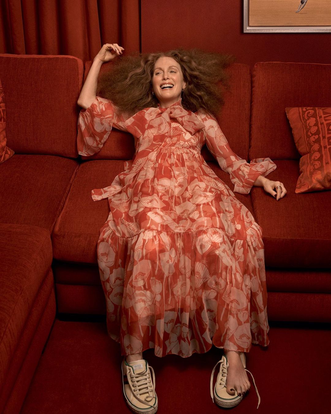 Woman in red floral dress lying on a red couch with white sneakers, smiling in an indoor setting with red walls and curtains.