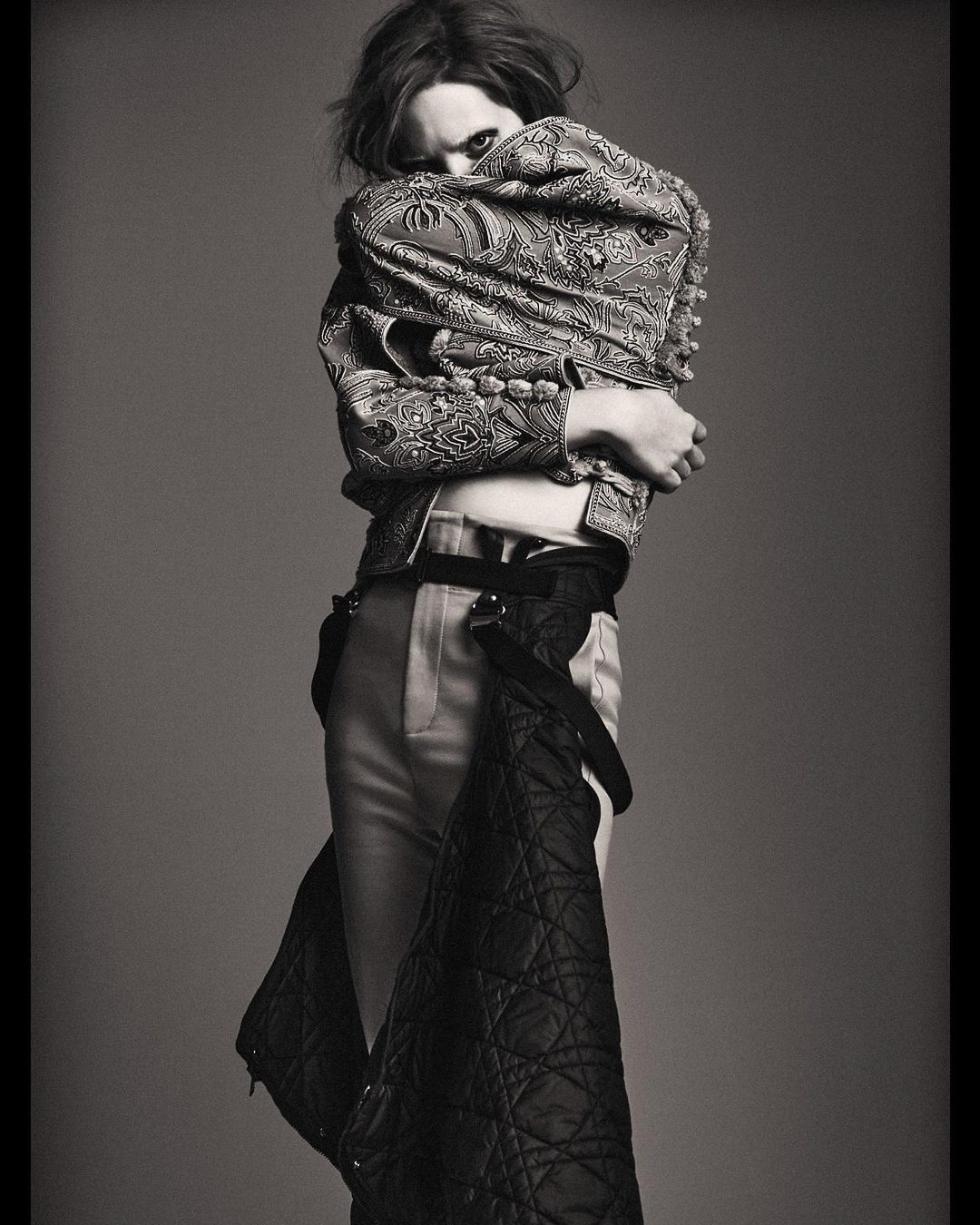 Monochrome fashion shot of an adult with medium hair, wearing a patterned jacket, light pants, and a quilted garment in a studio setting.