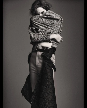 Monochrome fashion shot of an adult with medium hair, wearing a patterned jacket, light pants, and a quilted garment in a studio setting.