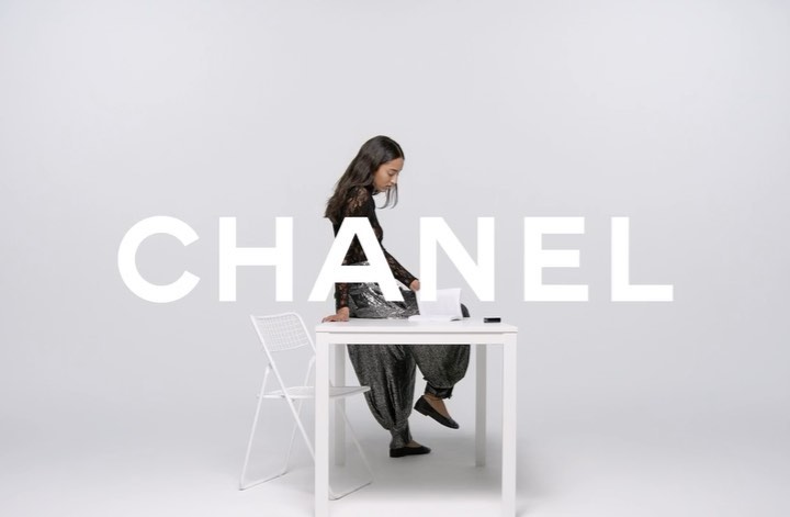 A young woman is sitting on a white table in a studio with white walls in a fashion photography setting, next to a white chair.