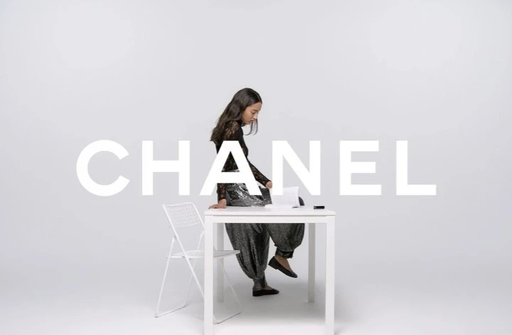 A young woman is sitting on a white table in a studio with white walls in a fashion photography setting, next to a white chair.
