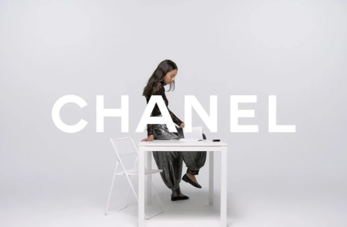 A young woman is sitting on a white table in a studio with white walls in a fashion photography setting, next to a white chair.