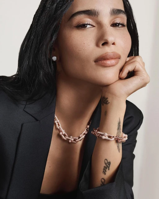 A close-up of a woman with dark hair wearing a black blazer, a light pink and silver chain necklace and bracelet, and a diamond earring.