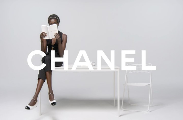 Fashion shot of a Black woman in a black outfit and white shoes seated on a white table, reading a book in front of a chair.