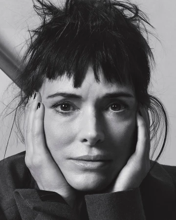 Monochrome portrait of a woman with dark hair and her hands on her face, wearing a dark-colored blazer against a neutral background.
