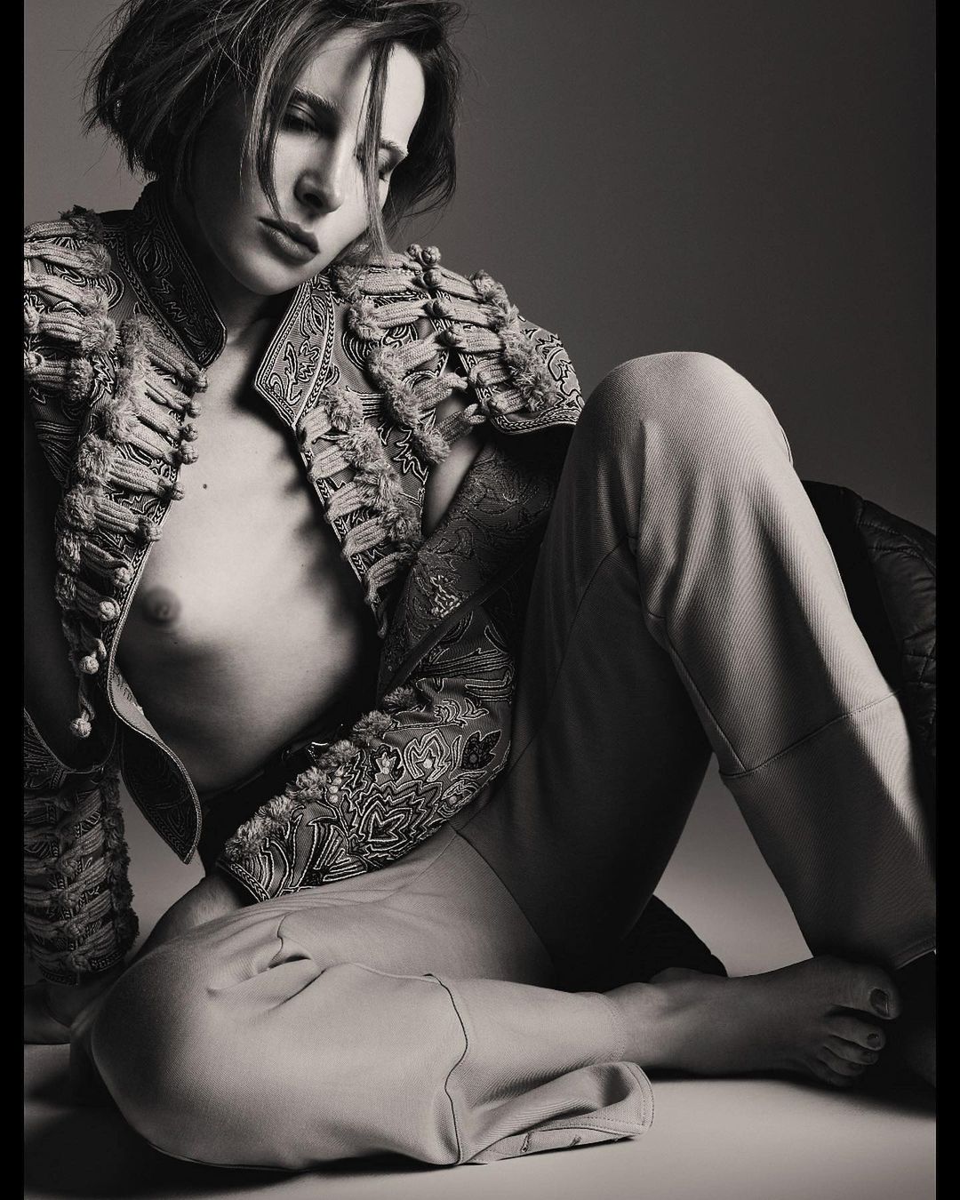 Monochrome studio portrait of a fair-skinned adult woman with short hair wearing an ornate jacket and trousers while seated.