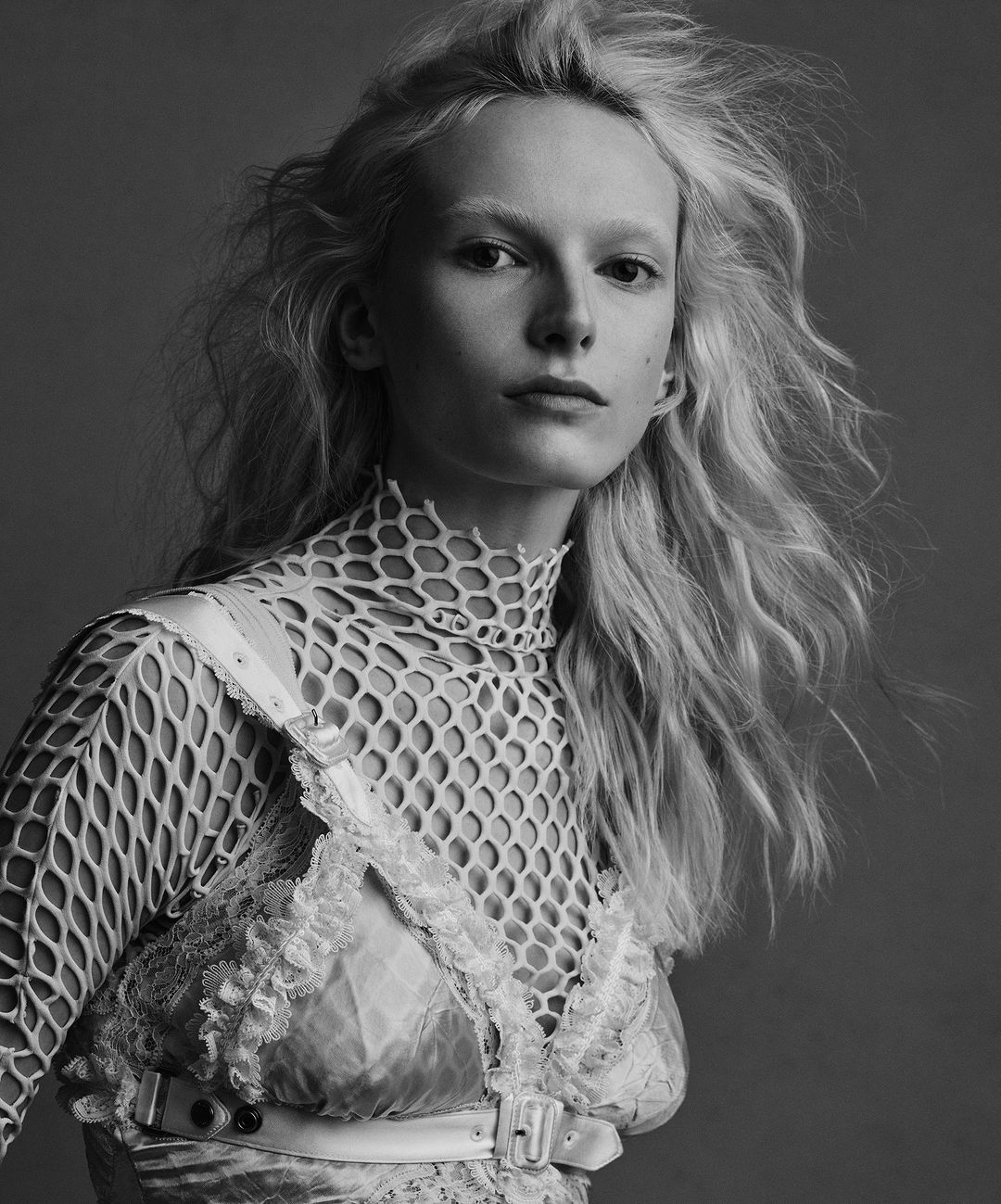 Monochrome portrait of a fair-skinned woman with blonde wavy hair, wearing a net top and lace dress against a grey backdrop in a photo studio.