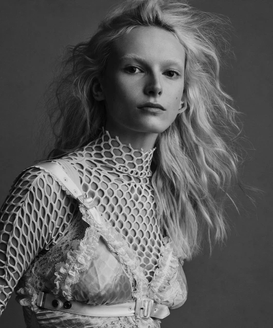 Monochrome portrait of a fair-skinned woman with blonde wavy hair, wearing a net top and lace dress against a grey backdrop in a photo studio.
