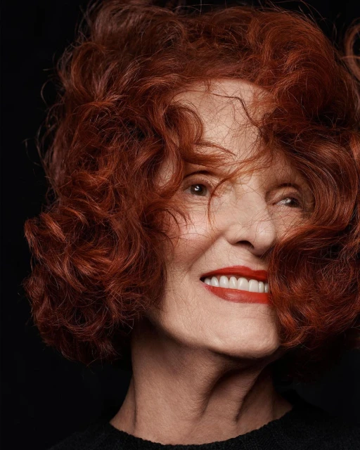 Close-up portrait of a smiling elderly woman with bright red curly hair and red lipstick against a dark, neutral background.
