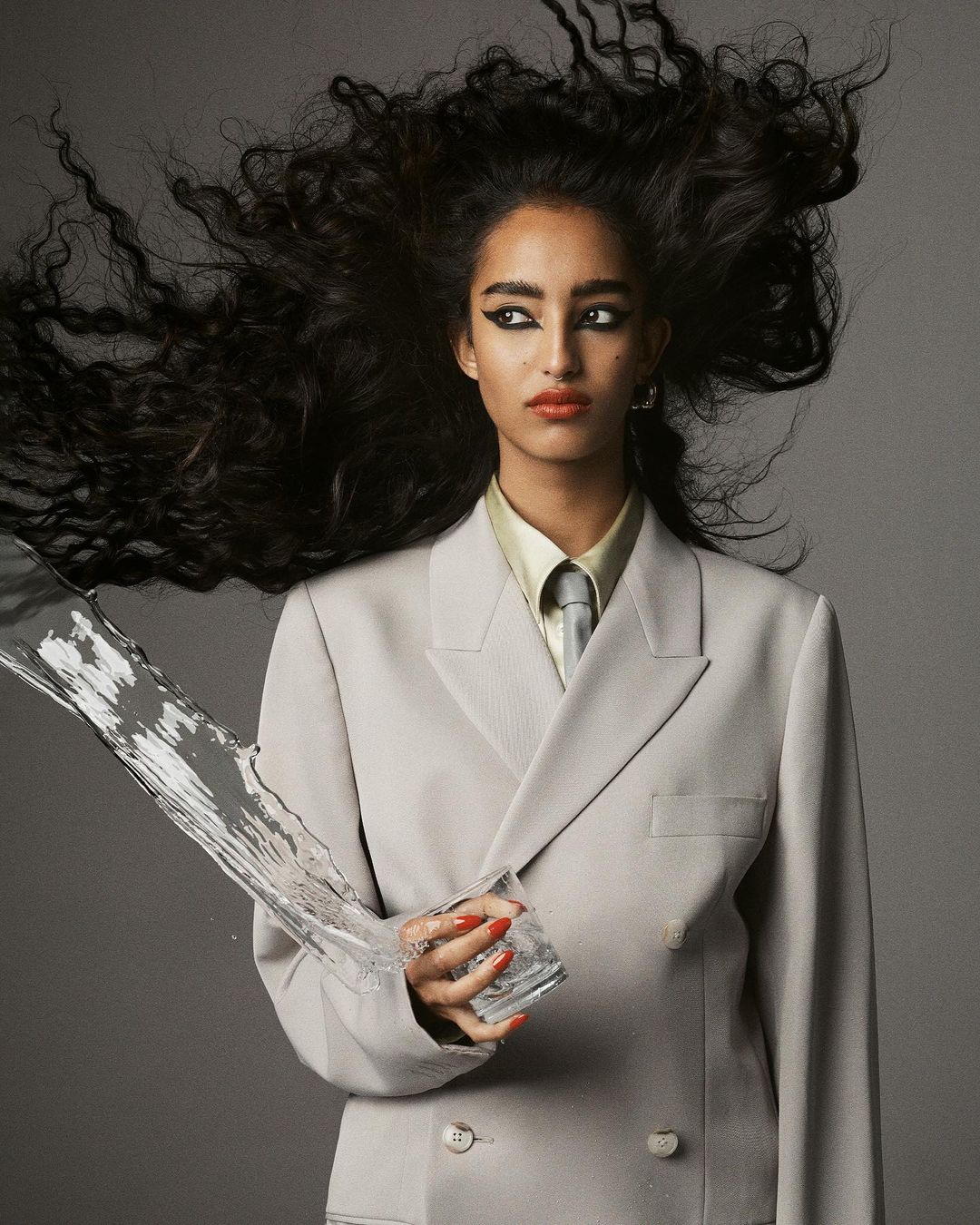 Glamorous woman in suit holding a glass of water that is splashing, with dark hair and makeup. Studio shot against gray background.