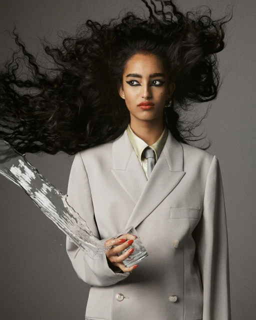 Glamorous woman in suit holding a glass of water that is splashing, with dark hair and makeup. Studio shot against gray background.