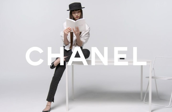 An Asian female model in a black hat and business casual attire sits on a white table while reading a book in a minimalist photo studio.