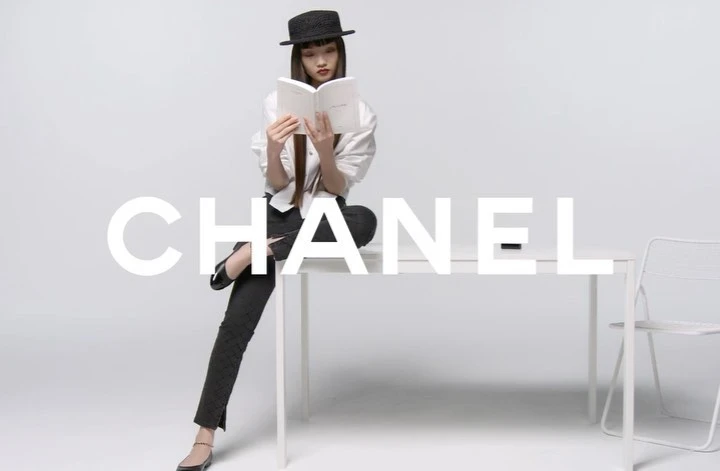 An Asian female model in a black hat and business casual attire sits on a white table while reading a book in a minimalist photo studio.