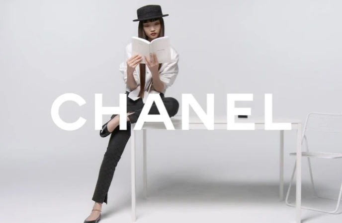 An Asian female model in a black hat and business casual attire sits on a white table while reading a book in a minimalist photo studio.
