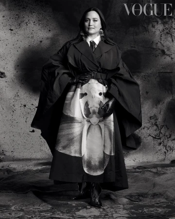 Lily Gladstone wearing a black coat and gloves standing in front of a textured wall