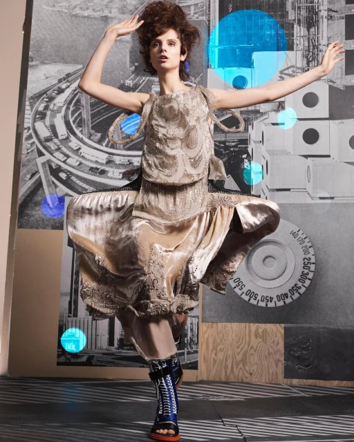 Fashion model in a vintage-style dress with elaborate lace-up boots, strikes a dance-like pose against an abstract, neutral-toned backdrop.