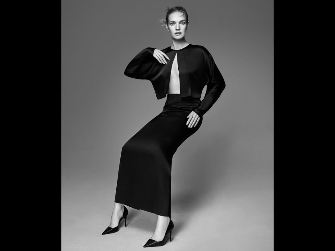 Fashion shot of a woman in a black two-piece outfit and high heels, posing against a grey background in a studio setting.